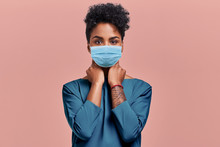 Gorgeous Dark Skinned Young Female With Afro Hairstyle Wearing Protective Mask Against COVID Virus, Tired Of Stress And Tension, Looks Confidently At The Camera, Poses Against Beige Studio Background