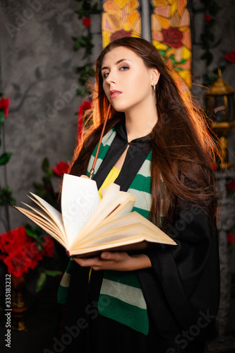 Fotografía A young woman in a black mantle with a striped scarf around her neck casts a spell from a book and conjures with a wand of magic