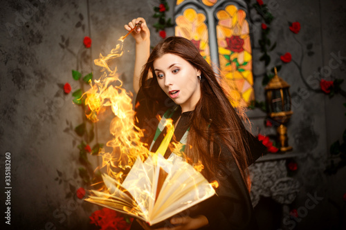 Cuadros en Lienzo A young woman in a black mantle with a striped scarf around her neck casts a spell from a book and conjures with a wand of magic