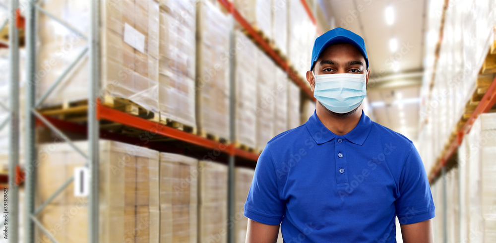 health protection, safety and pandemic concept - happy indian delivery man in blue uniform wearing face protective medical mask for protection from virus over warehouse background - obrazy, fototapety, plakaty 