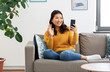 © Syda Productions - people, technology and communication concept - happy smiling asian young woman in yellow sweater sitting on sofa and having video call on smartphone at home