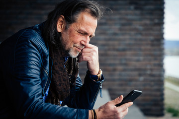 Older but cool man with beard and long grey hair uses a mobile phone in an urban environment