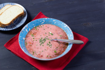 Canvas Print - Red beetroot soup borsch in blue plate with sour cream, spoon, green onion, bread  on textile cover on black wooden table