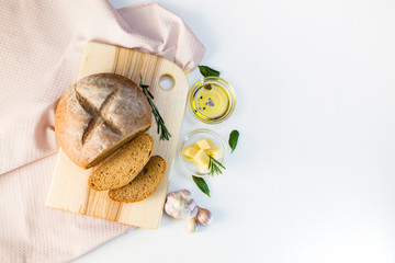 Round sliced bread with two slices lies on cutting board made of ash with a hole. White background with a light pink waffle towel, two bowls with olive oil and cheese, garlic, rosemary, spinach