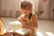 © sementsova321 - Close up portrait of cute toddler girl with dark hair reading book for her soft toy while sitting on floor in her bedroom, reads fairytale to her rabbit. Charming toddler in dress and with pigtails.