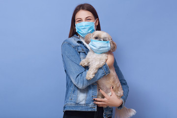 Indoor shot of woman dresses denim jacket protective flumask holds dog pet in medical mask. Coronavirus disease dangerous for people and pets. Coronavirus, pandemic, qaurantine, precautions concept.