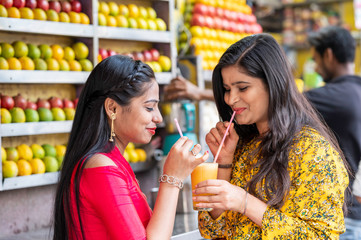 Young happy smiling Indian girls drinking or holding glass of orange juice outdoor in the market with colorful background of fruits. Urban healthy lifestyle.