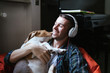 Young man hugs his beagle dog in his room while listening to music with headphones - Millennial is resting and having fun at home