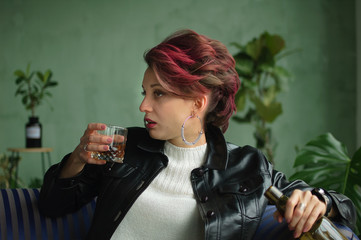 Beautiful sexy woman with dark pink hair in black leather jacket is drinking whisky from a glass sitting on blue sofa at home in loft style room, female alcoholism, alcohol addiction