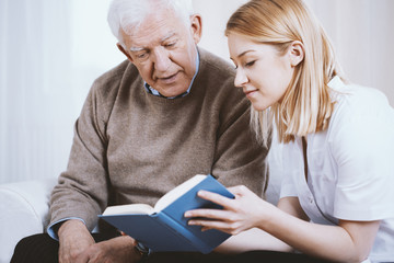 Young blonde volunteer reading book to senior man in nursing home