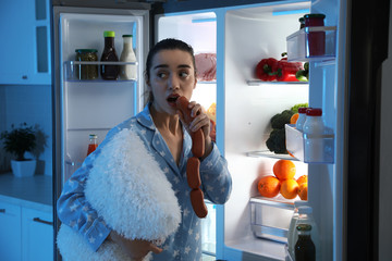 Poster - Young woman with pillow eating sausages near open refrigerator at night