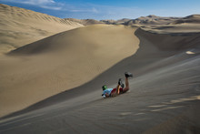 Man Sandboarding Down A Large Sand Dune In The Oasis Of Huacachina, Peru. Typical, Adventurous Day Activity In The Beautiful Pacific Coastal Desert With Views Of The Surreal Sandy Landscape