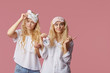Two twin sleepless young women in pajamas and sleep masks on a pink background. Alarm clock woke up the girls