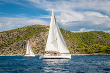 Two Sailing Yachts With White Sails Floating On The Sea Among The Rocks