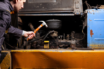 Wall Mural - An angry male mechanic is repairing the engine of an old truck with an open hood hitting it with a hammer.