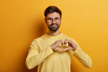 Handsome Bearded Adult Man Holds Hands In Shape Of Heart, Expresses His Affection To Girlfriend, Adores Someone, Says Be My Valentine, Dressed In Near Sweater, Isolated On Yellow Background.