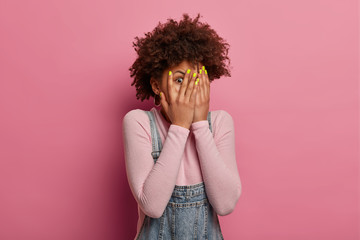 Wall Mural - Scared curly woman peeks through fingers, hides face with palms, afraids of terrifying scene as watches horror movie, sees phobia, stands alone, feels frightened, isolated on pink background