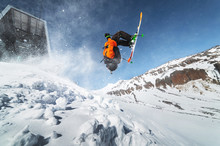 Low Angle View Athlete Skier In An Orange Jacket Does A Back Flip With Flying Powder Of Snow Against A Clear Blue Sky And Snow-capped Mountains Of The Caucasus.