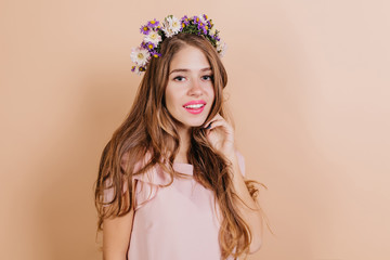 Wall Mural - Dreamy long-haired brunette woman with purple flowers in hair smiling to camera. Interested pretty girl standing on light background.