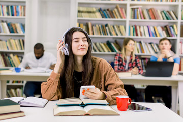 Pretty dreamy 25-aged brunette girl in casual shirt wearing earphones and enjoying favourite music during break after hard studying and preparation for exams in the college library