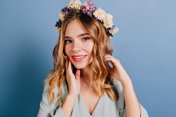 Wall Mural - Spectacular lady with pink lips and shiny hair posing on dark background. Adorable girl in flower wreath standing near blue wall.