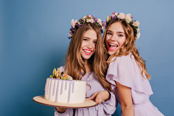 Wall Mural - Excited girls posing with cake and laughing on blue background. Elegant ladies celebrating something and holding big tasty dessert.