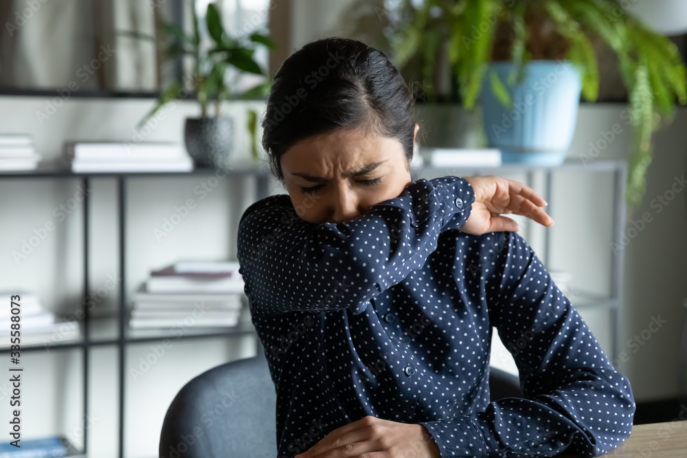 Millennial indian ethnic girl sitting in office, coughing in elbow, right illness behavior for not spreading virus infection. Unhealthy young woman feeling unwell at workplace, covid 19 symptoms. - obrazy, fototapety, plakaty 