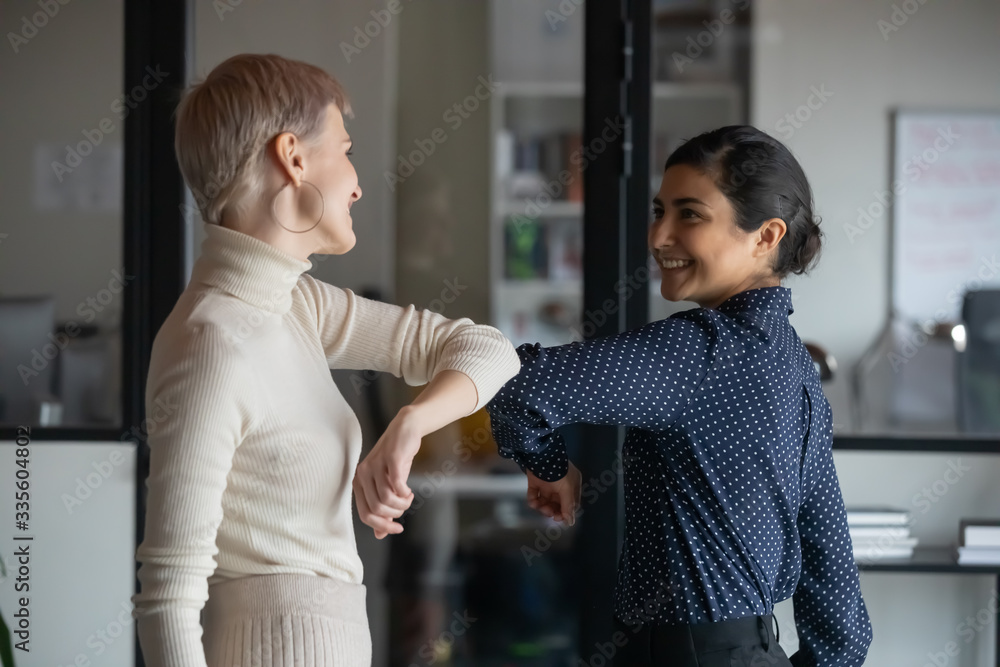 Friendly smiling millennial diverse female colleagues keeping social distance, greeting each other by bumping elbows instead of hugs or handshaking, preventing covid 19 coronavirus infection spread. - obrazy, fototapety, plakaty 