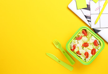 Fresh vegeterian salad in green lunch box with plastic fork and knife at office workplace.Close up of healthy snack in plastic container.Healthy food concept.Top view, flat lay,yellow background.