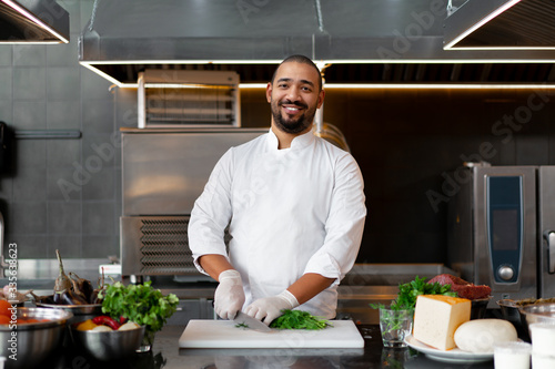 Fotomural Handsome young African chef standing in professional kitchen in restaurant preparing a meal of meat and cheese vegetables