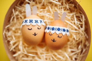 Two little easter eggs with a painted drawn rabbit faces and ears laying on the yellow table in hay isolated. Easter holidays decorations, preparations concept. Holy religious day. Copyspace.