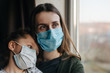 © Yura Yarema - Tired mother and little daughter in medical sterile face mask sitting on sill looking at window, sad family spending time alone at home. Concept of coronavirus or COVID-19 pandemic disease symptoms