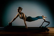 © Georgii - Young girl doing pilates exercises with a reformer bed. Beautiful slim fitness trainer on reformer gray background, low key, art light. Fitness concept