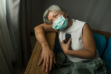 scared and worried attractive middle aged woman 50s with grey hair and protective mask during covid-19 virus crisis home lockdown quarantine sitting on couch  in fear