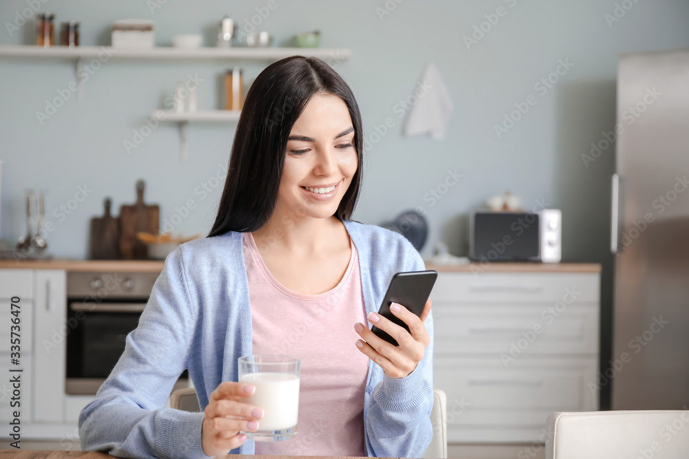 Beautiful young woman with mobile phone drinking milk in kitchen