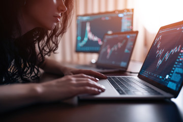 young female trader with laptop and screen looking at stock charts