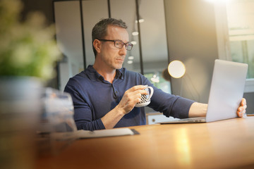 Wall Mural - Middle-aged guy having hot drink, working on laptop