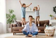 Happy Mother With Closed Eyes Meditating In Lotus Pose On Floor Trying To Save Inner Harmony While Excited Children Jumping On Sofa And Screaming In Light Spacious Living Room.