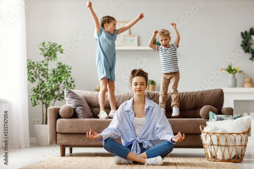 Fotomural Happy mother with closed eyes meditating in lotus pose on floor trying to save inner harmony while excited children jumping on sofa and screaming in light spacious living room