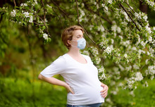 Portrait of a beautiful pregnant young woman wearing disposable medical face mask in the spring park during coronavirus outbreak.