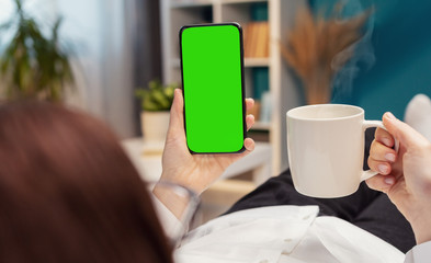 Rear view of person holding vertical cellphone in one hand and cup in the other lying on sofa in flat