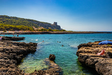 The Wonderful Bay Of Porto Selvaggio, With Pebbly And Rocky Beach. In Nardò, Italy, Puglia, Salento. People Sunbathe And Swim In The Turquoise Sea. The Torre Dell'Alto On The Promontory. Wild Nature.