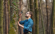 Half-body pic of focused botanist taking close look at ivy on tree standing in bare deciduous forest