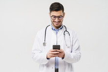 Young Male Doctor In White Coat Smiling While Looking At Screen Of His Phone, Using Medical App, Standing Isolated On Gray Background