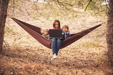 Working away from office concept, woman with two kids working on laptop while lying in hammock in a forest.