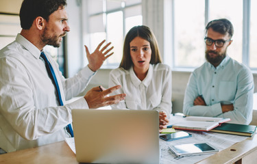 Tired office workers feeling headache during deadline reading email with bad news from colleague about project, unhappy male and female partners near desktop with blueprint and modern laptop computer