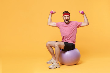 Wall Mural - Cheerful bearded fitness sporty guy sportsman in headband t-shirt in home gym isolated on yellow background. Workout sport motivation lifestyle concept. Sit on fitball, doing exercise with dumbbell.