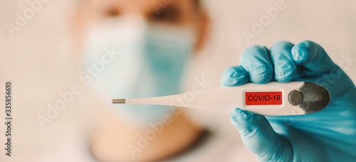 Closeup man in medical face mask and glove showing digital thermometer with COVID-19 text. Doctor in protective glove and mask holding electronic thermometer in hand. Coronavirus symptoms, diagnosis
