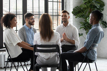 Wall Mural - Happy handsome male mentor counselling speaking with diverse people sitting in circle at group therapy session. Business coach training staff, having fun, team building activity at work.