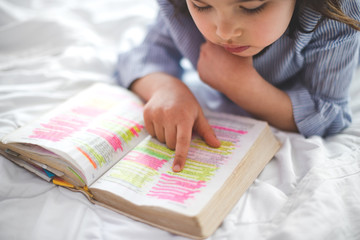 Wall Mural - Little girl reading from Bible while lying in bed. Bible with marks on letters.